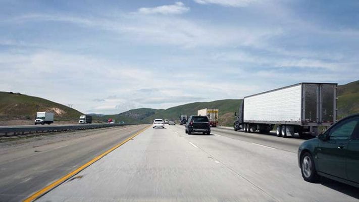 Cars passing big rig trucks on California highway, with green hills in background.